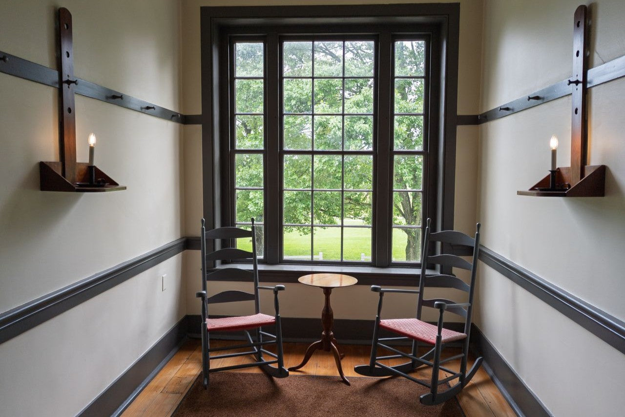 Cozy Shaker room featuring rocking chairs and candle sconces by a window. Perfect for serene moments.