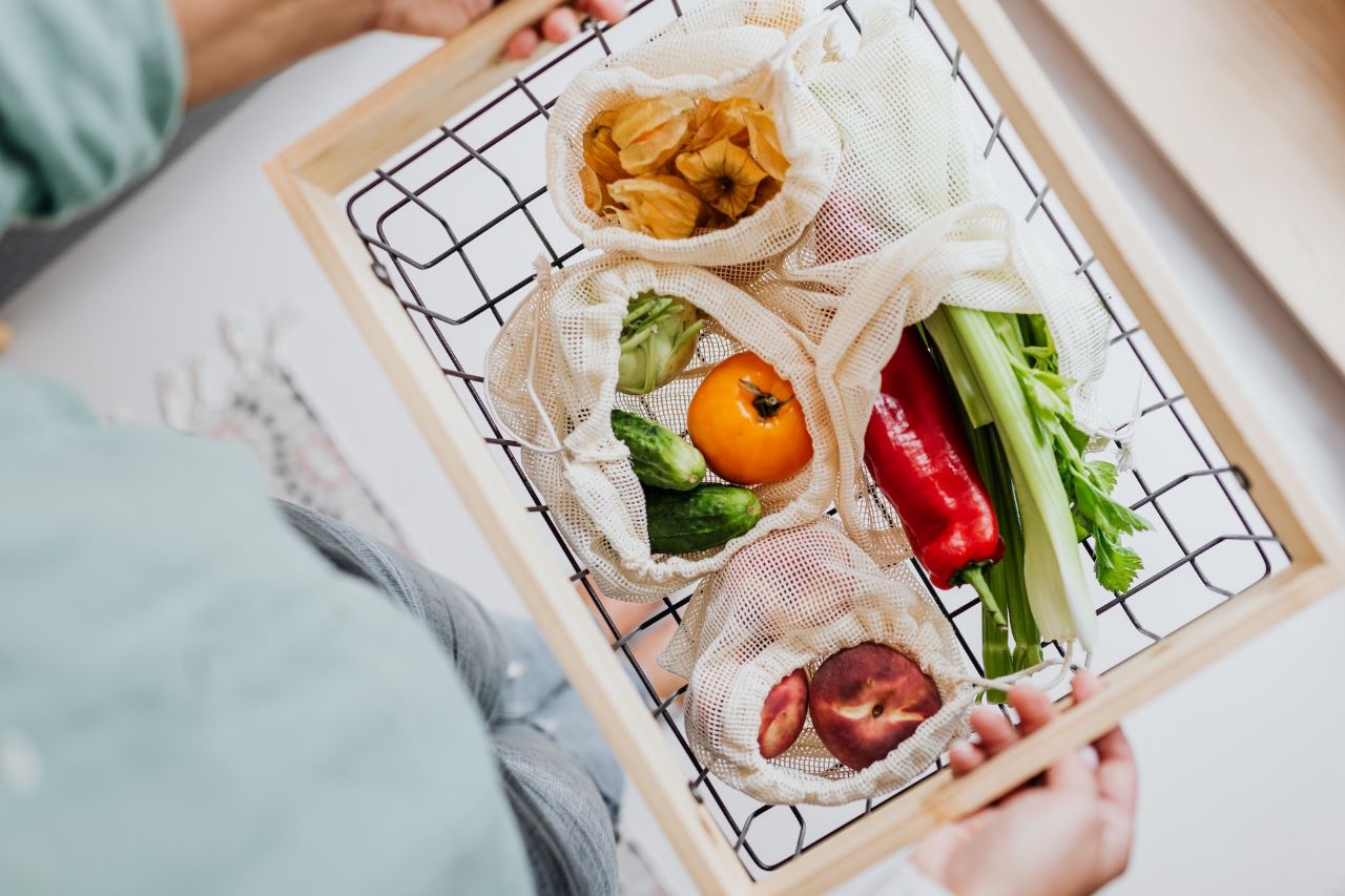 Person carrying a basket of fruits and vegetables that are wrapped in reusable bags