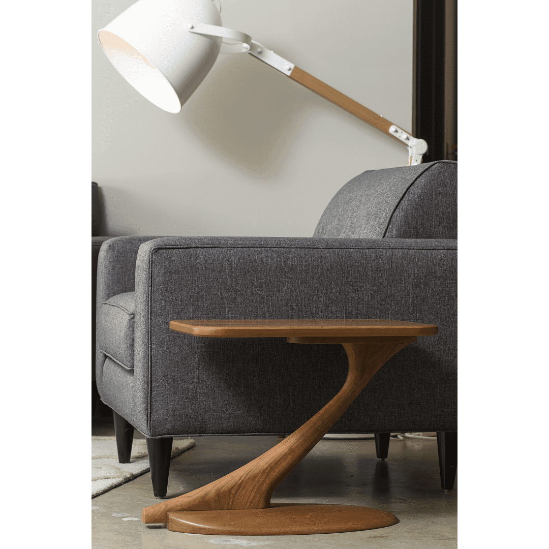 A modern living room featuring a gray upholstered sofa, the TY Fine Furniture Cantilever Side Table Solid Wood Handmade in Cherry with a curved base, and a large white floor lamp with a wooden arm behind the sofa.