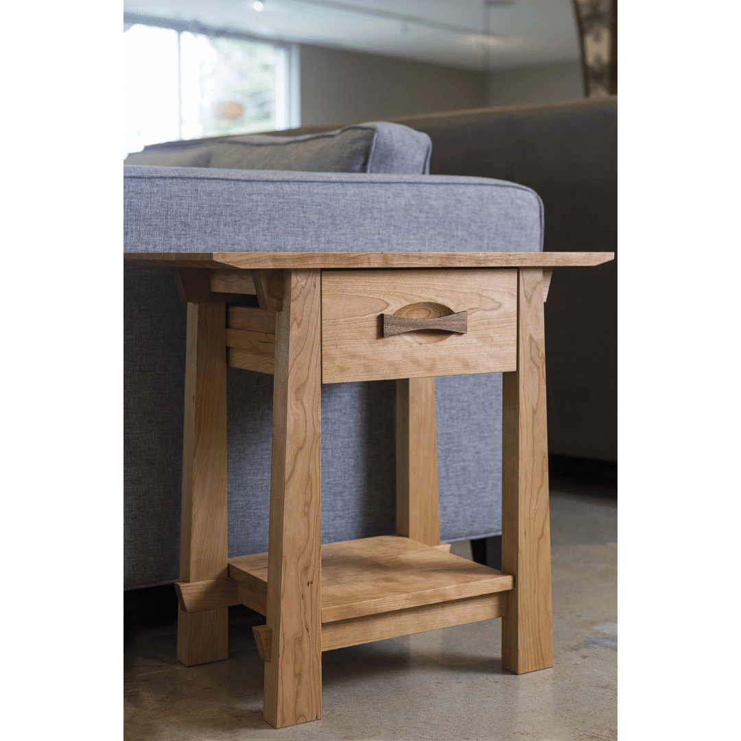 The Enso Side Table With Drawer by TY Fine Furniture, a handmade solid wood piece, sits beside a gray sofa in a modern living room with concrete floors, offering organic style plus convenient storage with its drawer and lower shelf.