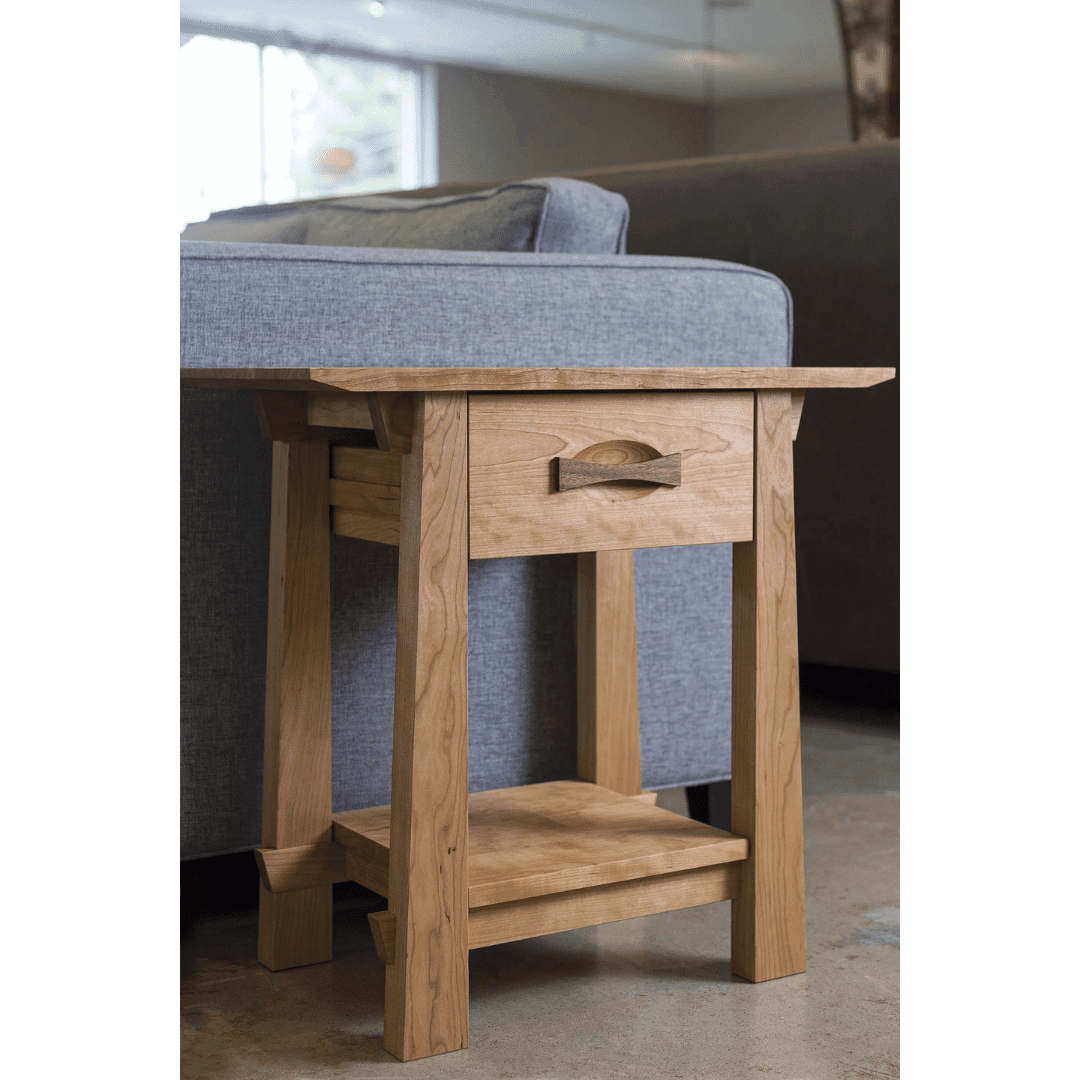 The Enso Side Table With Drawer Solid Wood Handmade Organic- Cherry by TY Fine Furniture sits beside a gray upholstered sofa in a living room with a concrete floor, offering storage with its drawer and lower shelf.