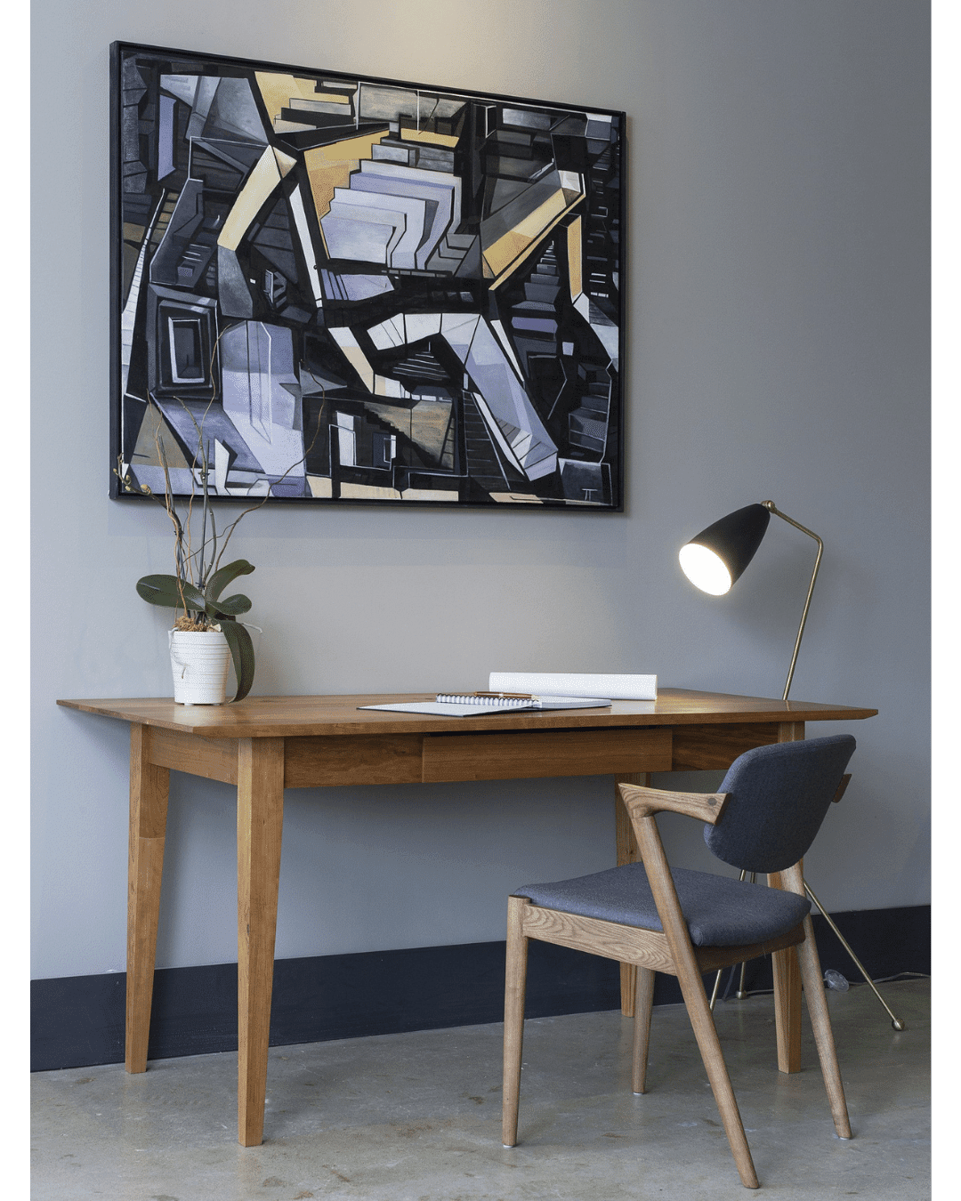 The Enso Desk by TY Fine Furniture, a solid wood handmade piece with cable grommets, stands against a gray wall with a matching chair, desk lamp, potted orchid, and an abstract black, gray, and yellow geometric painting overhead.