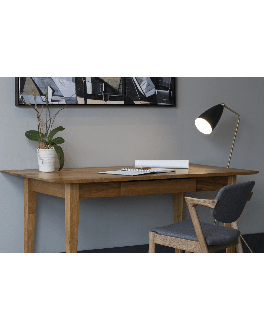 The Enso Desk by TY Fine Furniture features solid wood artisan craftsmanship, built-in cable grommets for tidy wire management, and pairs perfectly with a gray cushioned chair, potted plant, notebook, and modern black lamp under framed art.