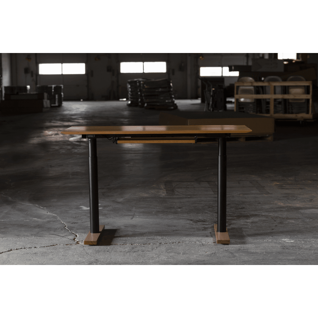 A TY Fine Furniture Enso Sit-Stand Desk with a solid wood top and black adjustable legs stands in the center of a dimly lit industrial warehouse with concrete floors and equipment stacked in the background.