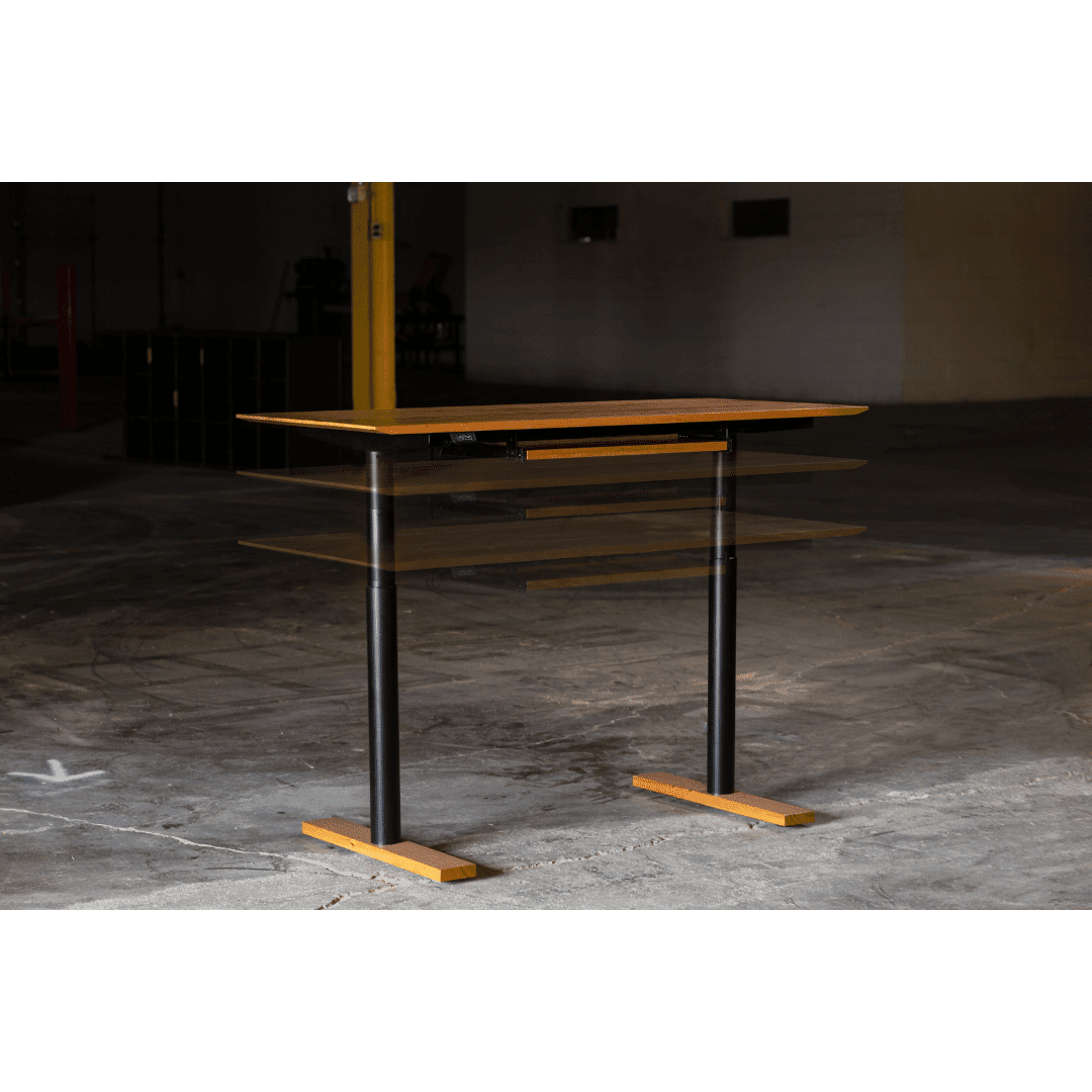 A TY Fine Furniture Enso Sit-Stand Desk - Solid Wood Top | Columbus OH with a black frame is displayed at three heights in an empty industrial space with concrete floors and a yellow post.