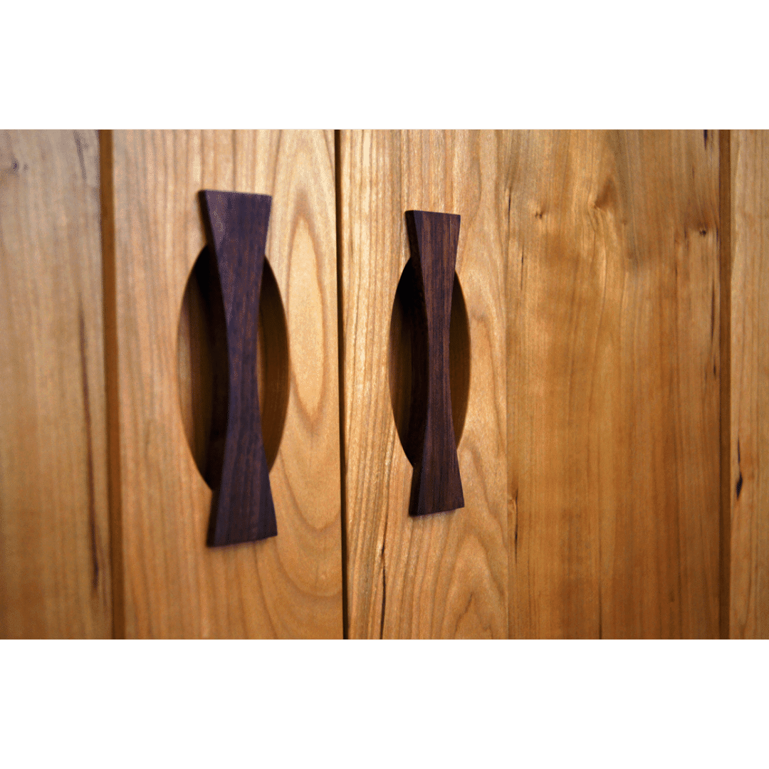Close-up of two wooden cabinet doors on the Enso Armoire by TY Fine Furniture, Columbus OH—solid hardwood with bowtie-shaped dark wood handles and oval cutout backplates on a light surface, matching its adjustable shelves.