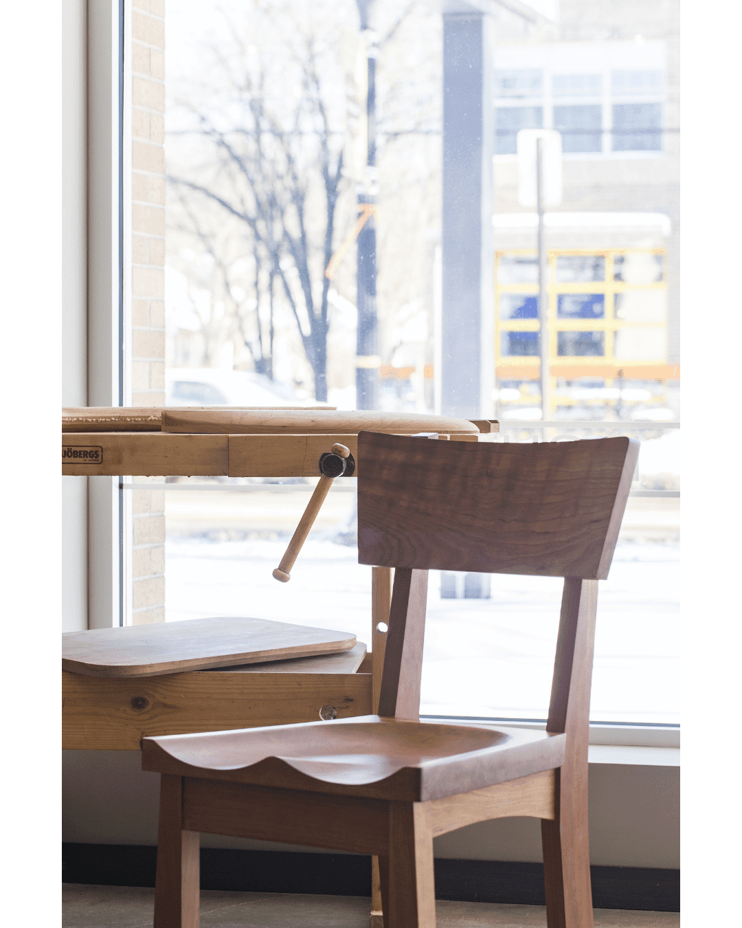 A TY Fine Furniture Enso Dining Chair—solid wood and handcrafted in Columbus, Ohio—is set before a large window beside a workbench with a metal crank, with snowy scenery and tree-lined streets evoking Japanese woodworking serenity.