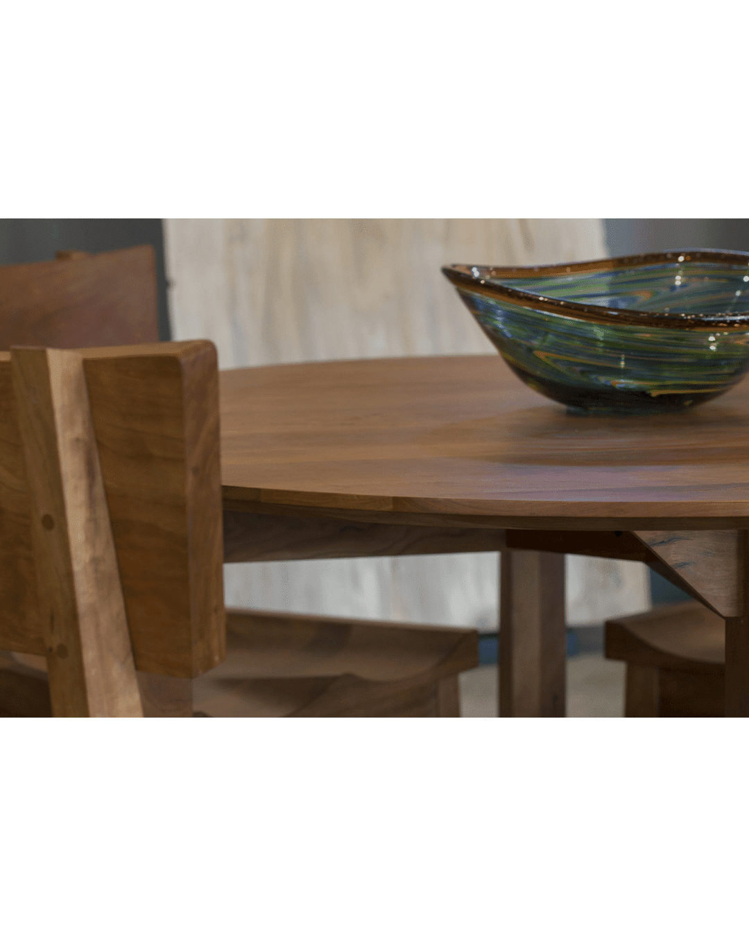 A round wooden table with TY Fine Furniture’s Enso Dining Chairs—solid wood and handcrafted in Columbus, Ohio—paired with a decorative glass bowl accented in green and blue, blending contemporary appeal with classic elegance.