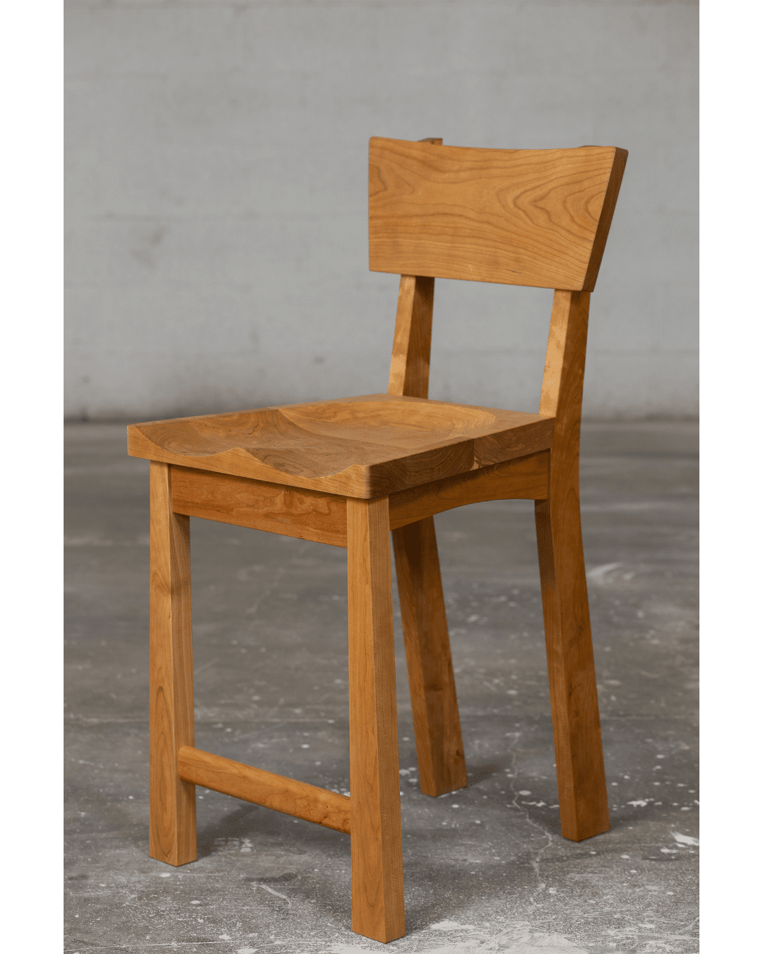 The Enso Barstool - Bar & Counter Height by TY Fine Furniture features a flat seat, straight legs, and a slightly angled backrest, standing on a concrete floor against a plain gray wall in Columbus OH.