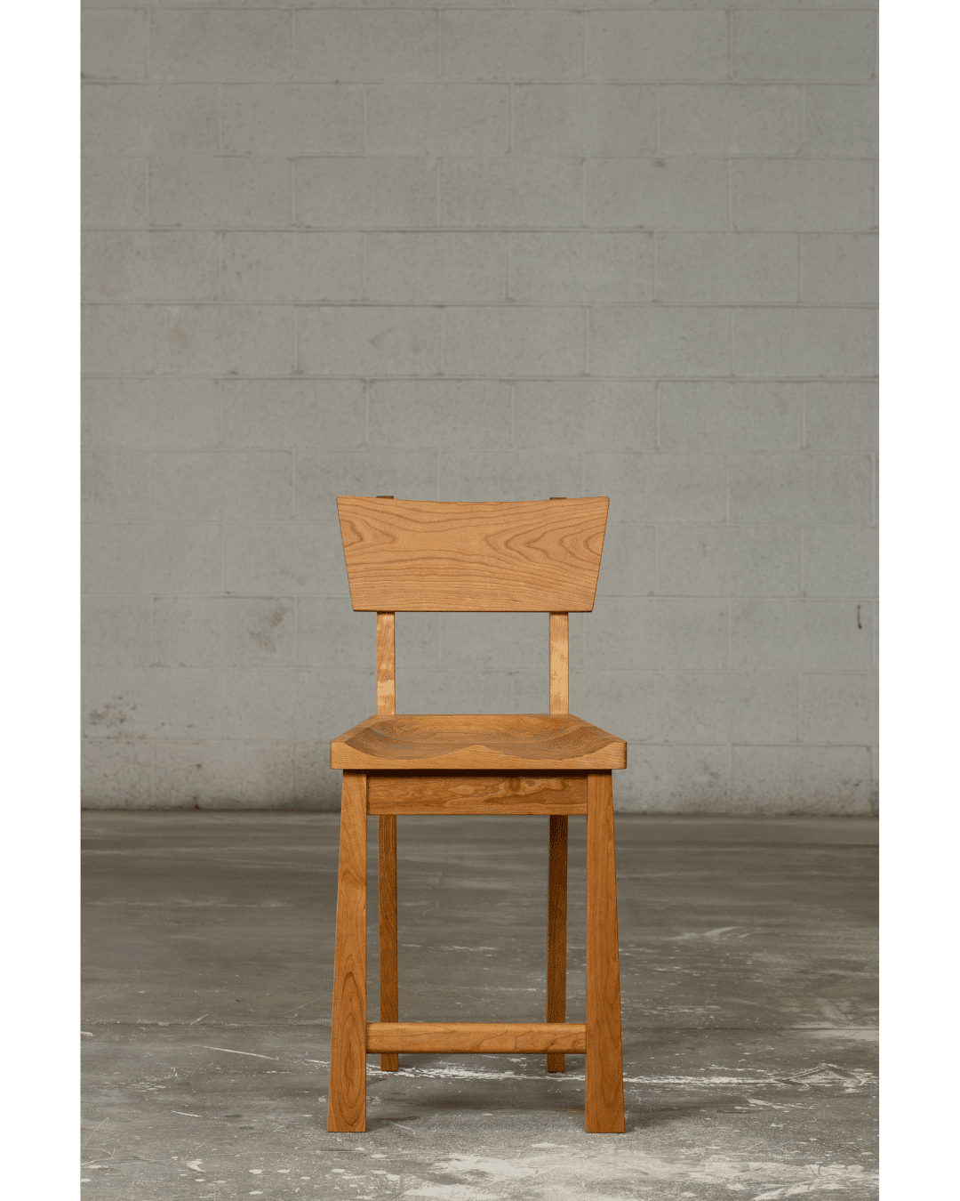 The Enso Barstool by TY Fine Furniture features a simple solid wood design with a flat seat and angular backrest, shown on a concrete floor against a plain gray brick wall. Available in bar and counter height in Columbus, OH.