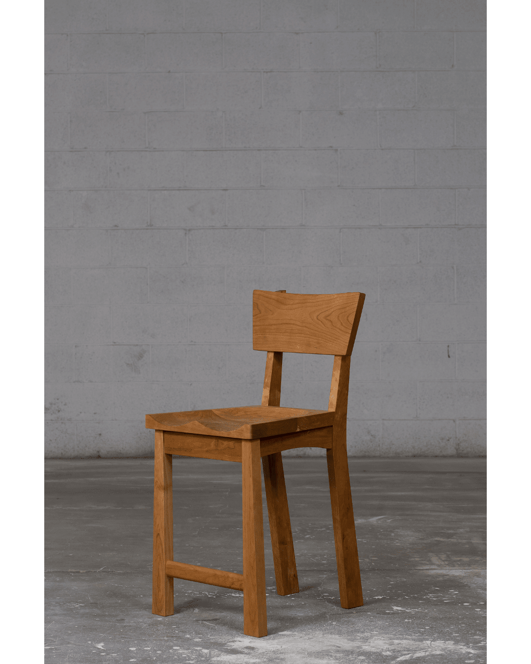 The Enso Barstool by TY Fine Furniture features a solid wood frame, straight backrest, and square seat, shown on a concrete floor against a plain gray brick wall. Available in bar and counter height in Columbus, OH.