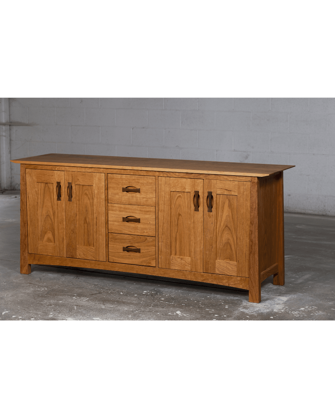 The Enso Solid Wood Buffet by TY Fine Furniture features four cabinet doors and three central drawers, offering stylish dining room storage. Shown in a natural finish, it stands on a concrete floor against a plain gray wall.