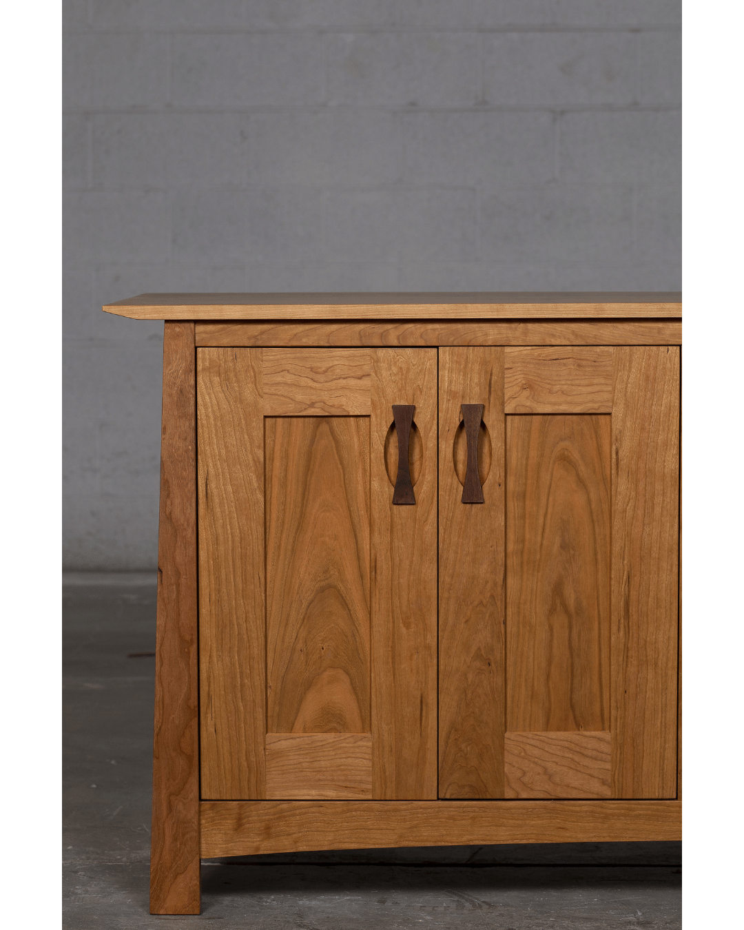 The Enso Solid Wood Credenza Buffet by TY Fine Furniture, handmade from cherry, features two paneled doors with dark handles. Only its left side is visible as it stands on a concrete floor against a plain gray wall—ideal for dining room storage.