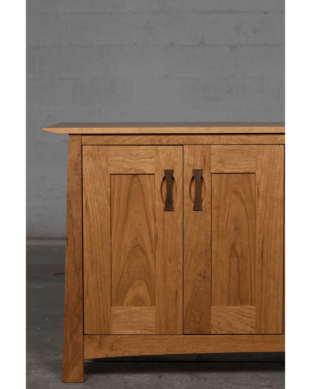 The left half of the TY Fine Furniture Enso Solid Wood Buffet, featuring two paneled doors and curved handles, is shown against a plain gray concrete wall and floor. This sustainable dining room storage piece is available in Columbus, OH.