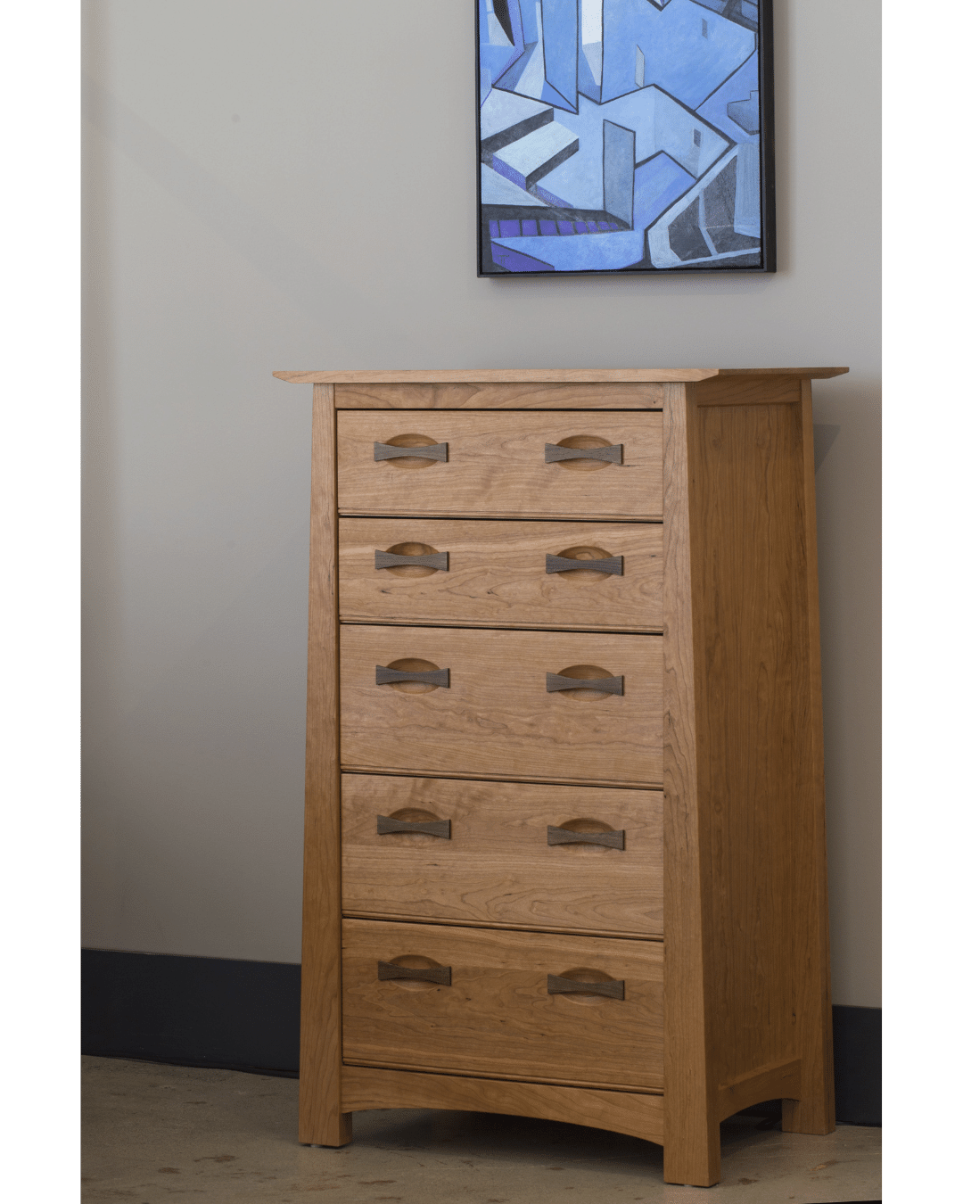 The Enso 5-Drawer Upright Dresser by TY Fine Furniture is a handmade solid cherry wood chest with sleek metal handles, perfect for bedroom storage and stylishly set beneath abstract blue artwork against a light wall.