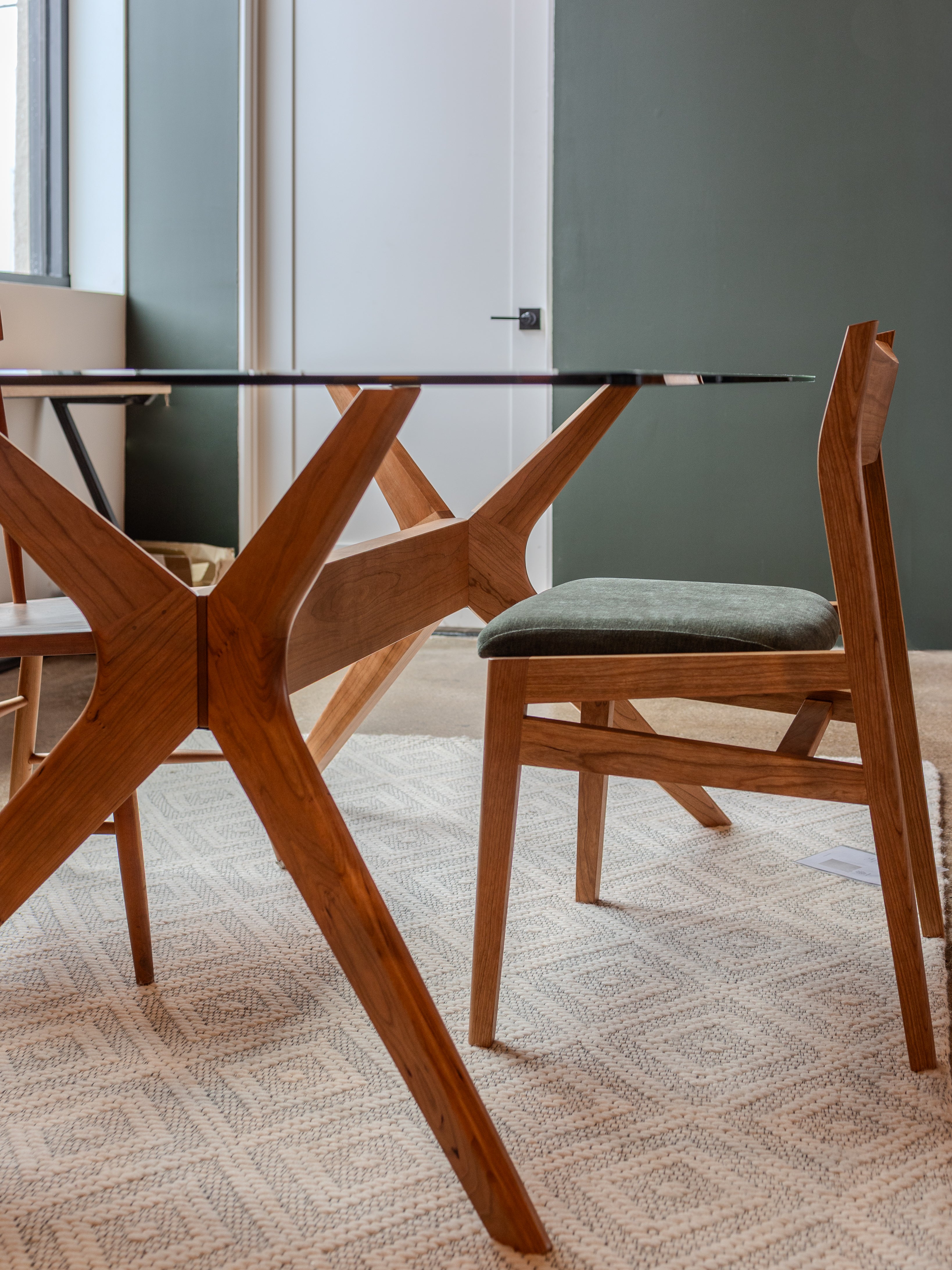 The Solid Cherry Dining Chair from the Strata Collection by T.Y. Fine Furniture, featuring a green cushion, is placed beside a glass table with wooden legs on a patterned rug in a stylish room with green and white walls.