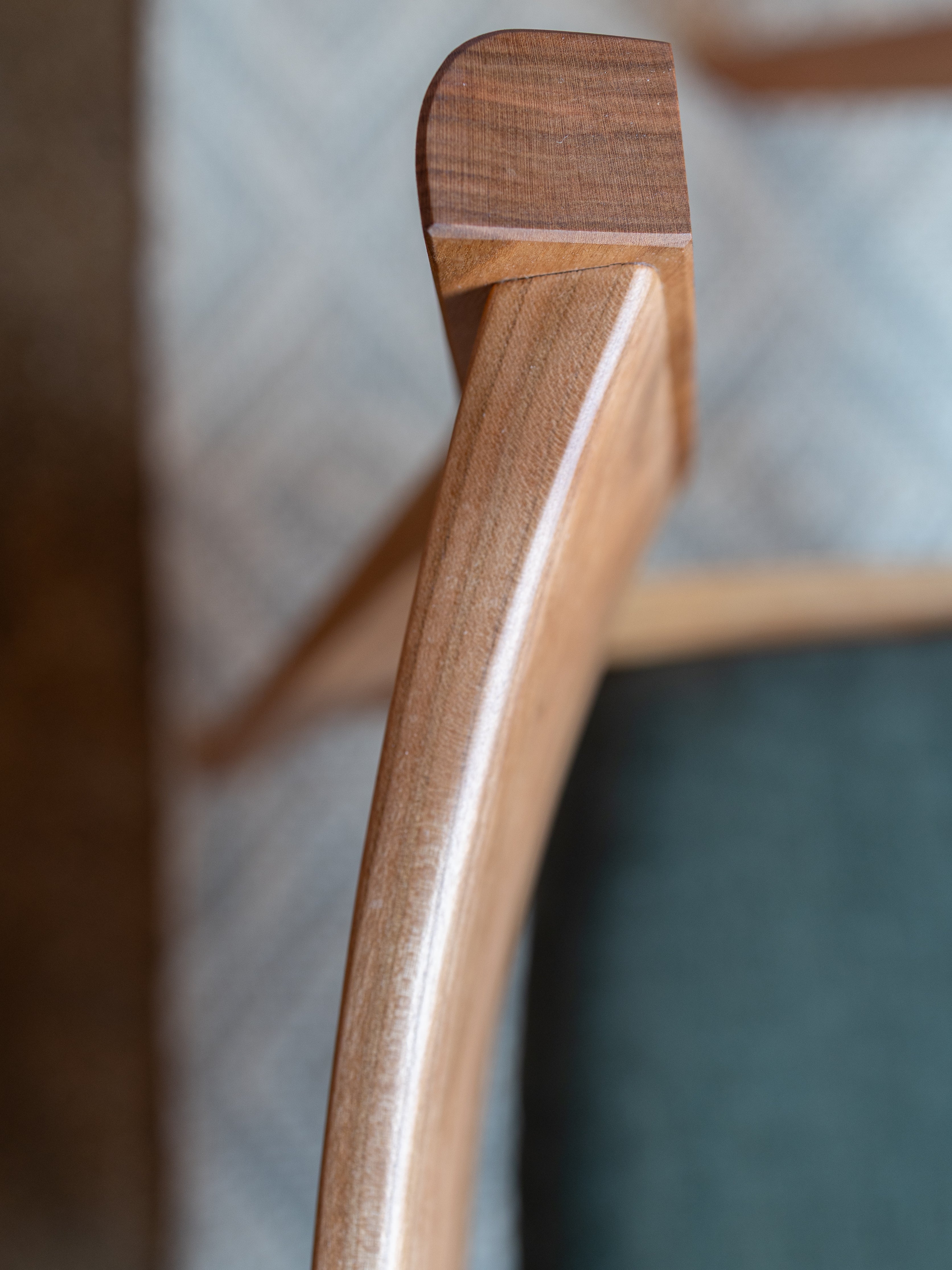Close-up of the Solid Cherry Dining Chair - Strata Collection by T.Y. Fine Furniture, highlighting its handcrafted armrest with smooth, polished cherry wood grain and an organic finish, set against a backdrop of blurred patterned fabric.