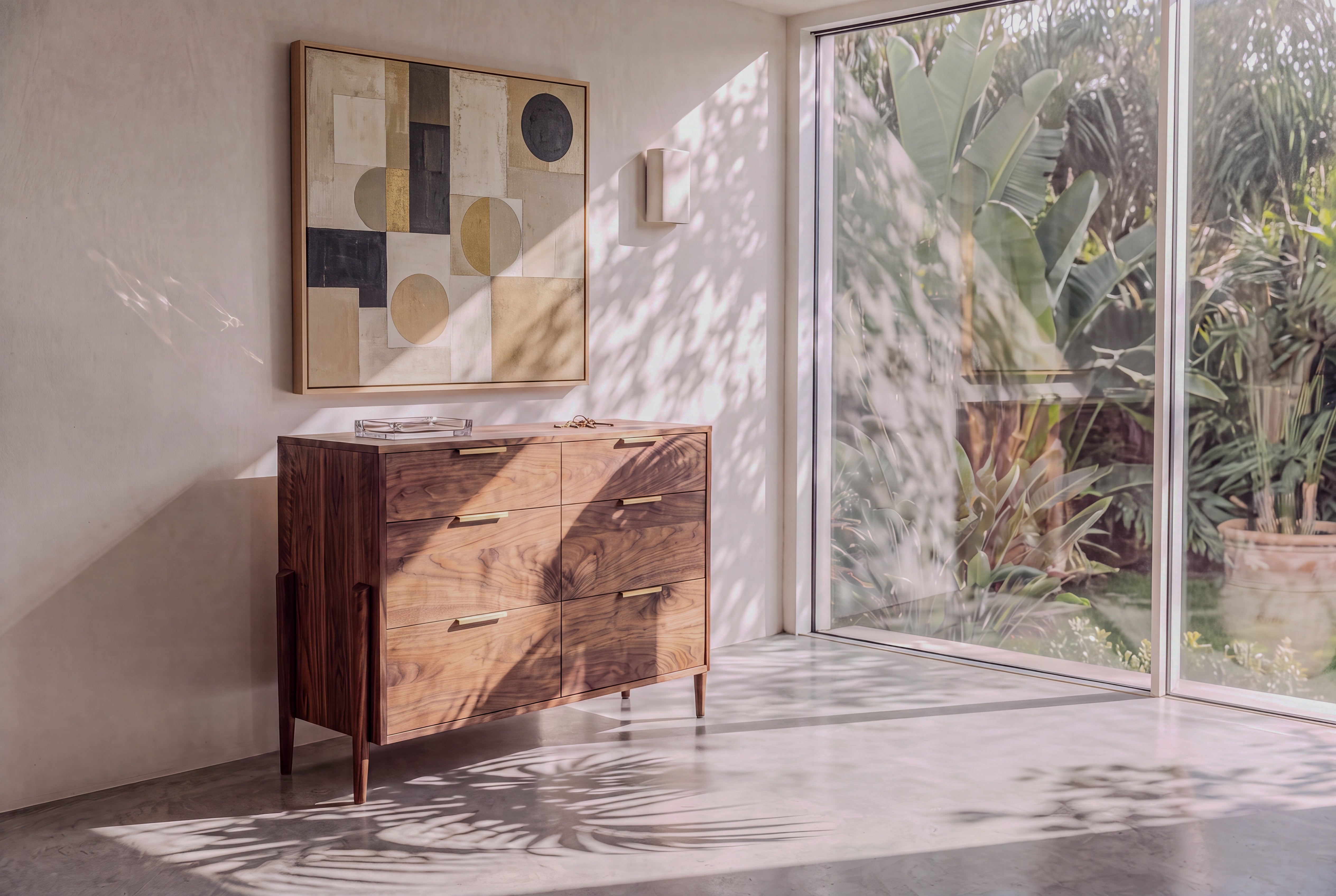 A Solid Cherry 6-Drawer Double Bank Dresser from the Apollo Collection by TY Fine Furniture stands against a white wall beneath abstract art. Sunlight pours through large glass windows, casting leaf shadows on the floor, while lush green plants flourish outside.