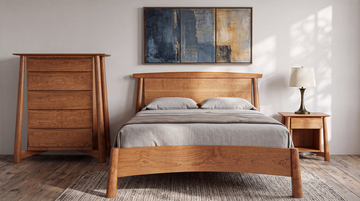 A wooden bed frame, matching heirloom-quality Sunrise Side Table - Cherry, and chest of drawers set in a bedroom with a table lamp and wall art.