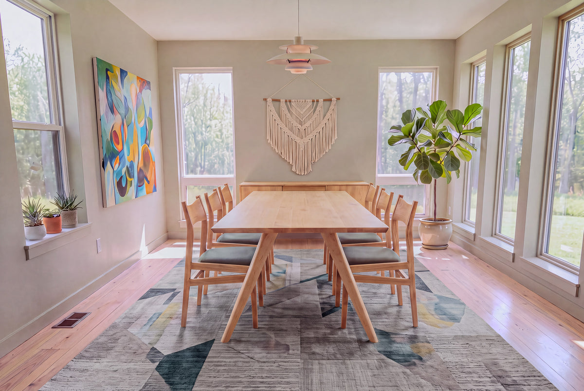 Bright dining room with large windows, a Solid Maple Dining Table from the Essentials Collection by TY Fine Furniture surrounded by wooden chairs, abstract painting, macramé wall art, potted plants, and a geometric rug on light wood floor.