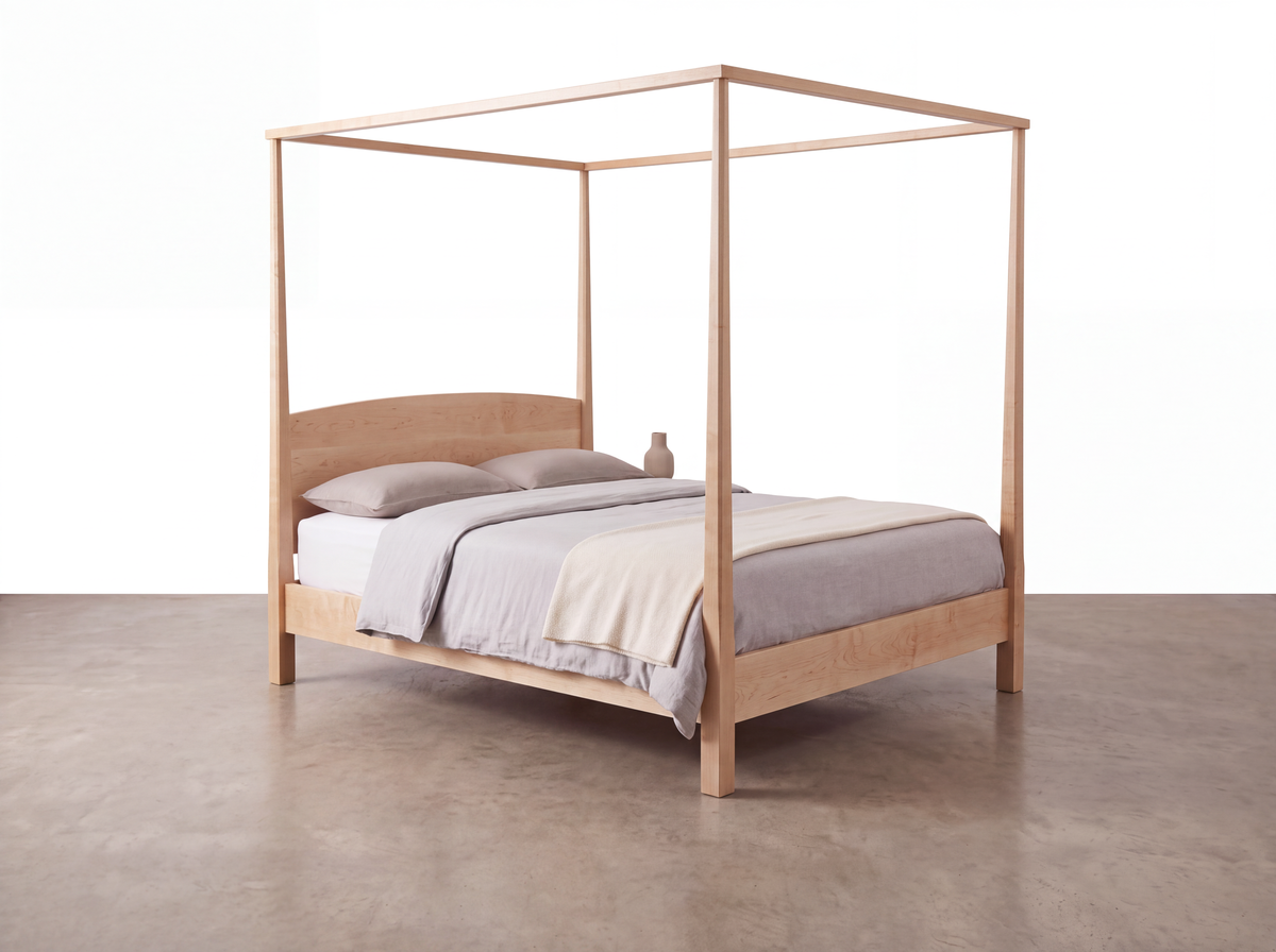 A Solid Maple Pencil Post Canopy Bed from the Shaker Collection by TY Fine Furniture, featuring light gray bedding, two pillows, a beige throw blanket, and a small vase placed on the right, all set atop a smooth concrete floor against a plain white background.