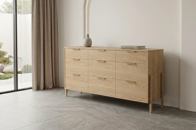 A contemporary Solid White Oak 9-Drawer Triple Bank Dresser from the Apollo Collection by TY Fine Furniture, featuring sleek gold handles and adorned with a small vase and a book on top, stands against a light-colored wall near expansive windows draped with beige curtains.