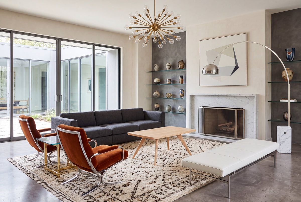 Modern living room with a gray sofa, two orange chairs, a white bench, the Solid Maple Square Coffee Table from TY Fine Furniture’s Essentials Collection, a patterned rug, fireplace, geometric wall art, and glass shelves displaying ceramics. Large windows let in natural light.