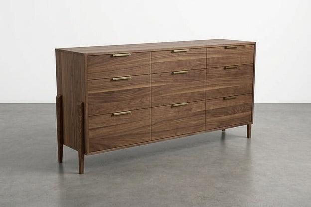 The Solid Walnut 9-Drawer Triple Bank Dresser from the Apollo Collection by TY Fine Furniture—a mid-century modern piece featuring brass handles—stands on four legs in an empty room with gray flooring and a white wall.