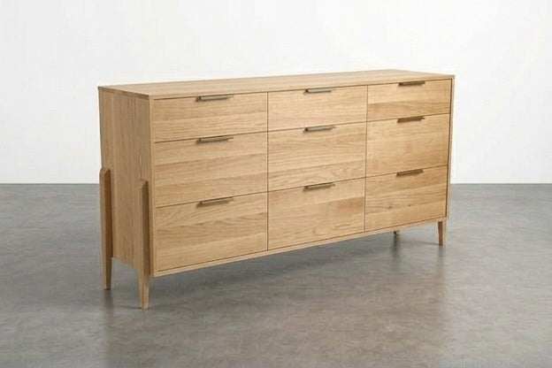 A light solid white oak 9-drawer triple bank dresser from the Apollo Collection by TY Fine Furniture stands on angled legs in a minimalist room with a gray floor and white wall, its simple metal handles and refined design embodying timeless elegance.