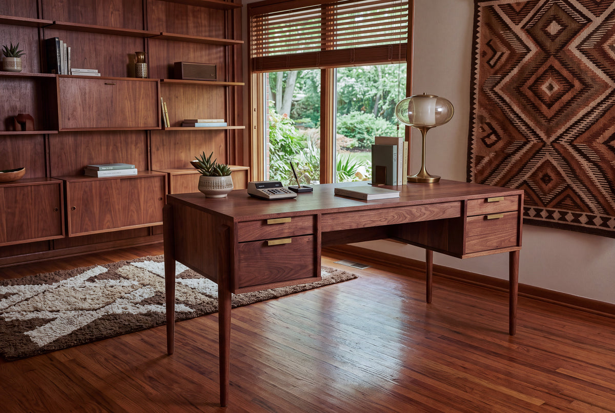 A mid-century modern office featuring the Solid Walnut Executive Desk - Apollo Collection | TY Fine Furniture, plus a lamp, books, plants, shelves, rug, and a large window with greenery outside.