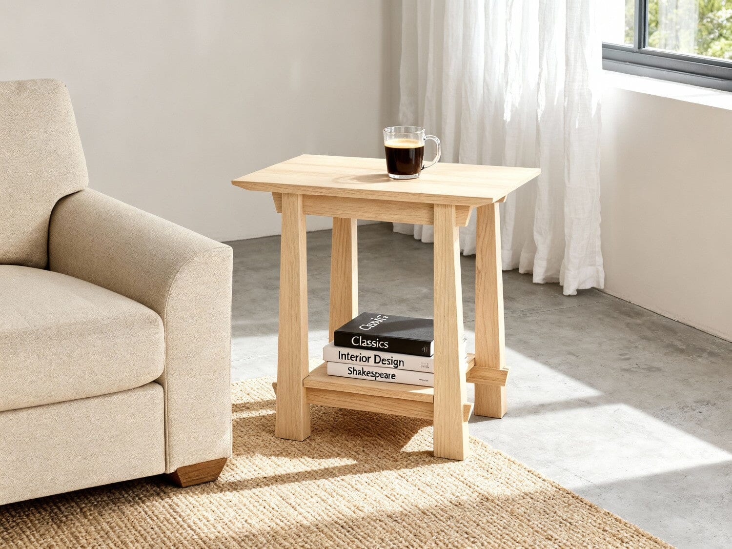 The Horizon Side Table in Maple by TY Fine Furniture sits beside a beige sofa, topped with a glass mug of coffee. Sunlight filters through white curtains onto a woven rug and concrete floor. Three books are stacked neatly on its lower shelf.