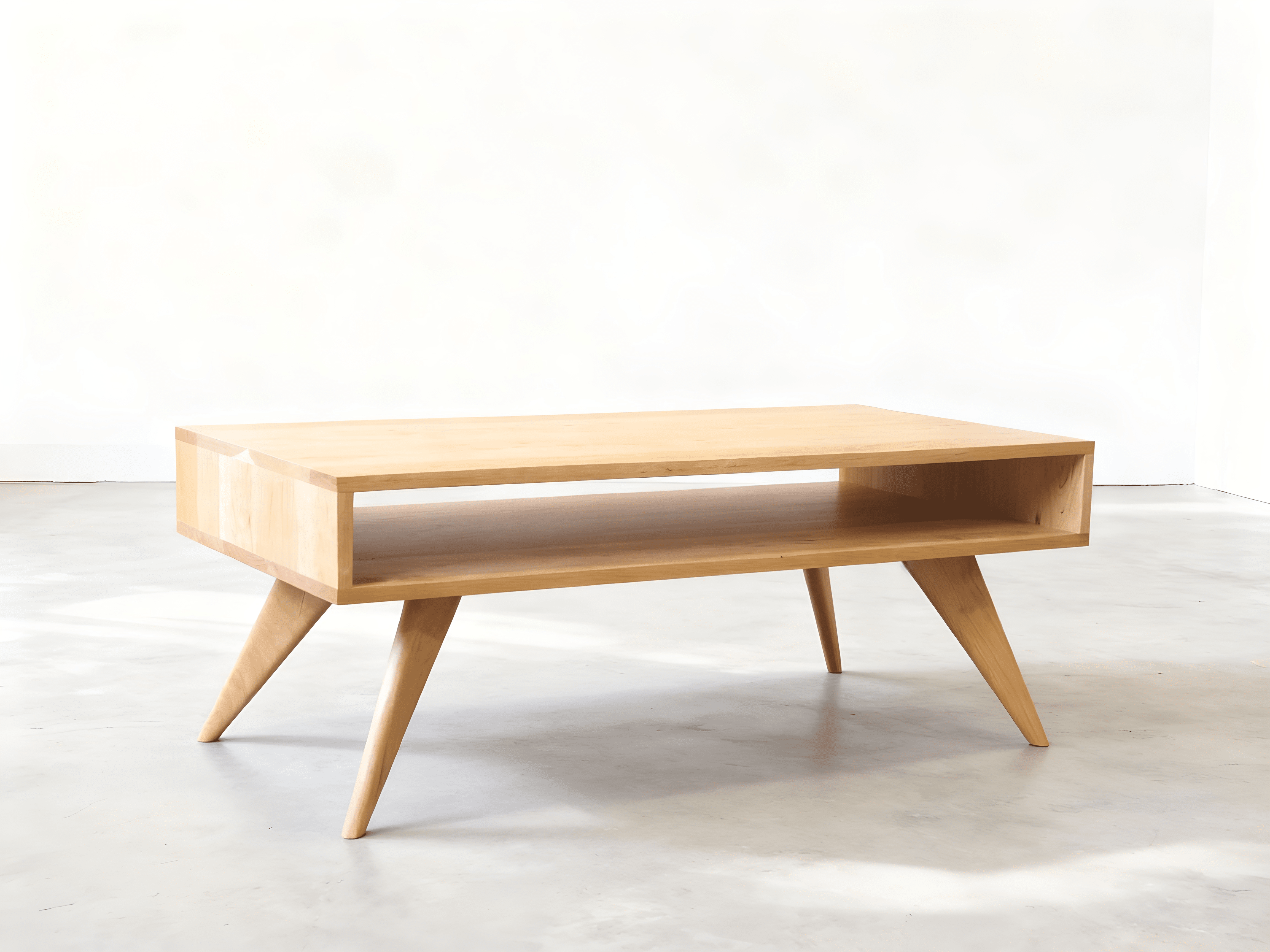 The TY Fine Furniture Modern Coffee Table in Maple - Solid Wood features angled legs and an open shelf, standing on a smooth light gray floor in a bright, minimalist room. Available in Columbus, OH.