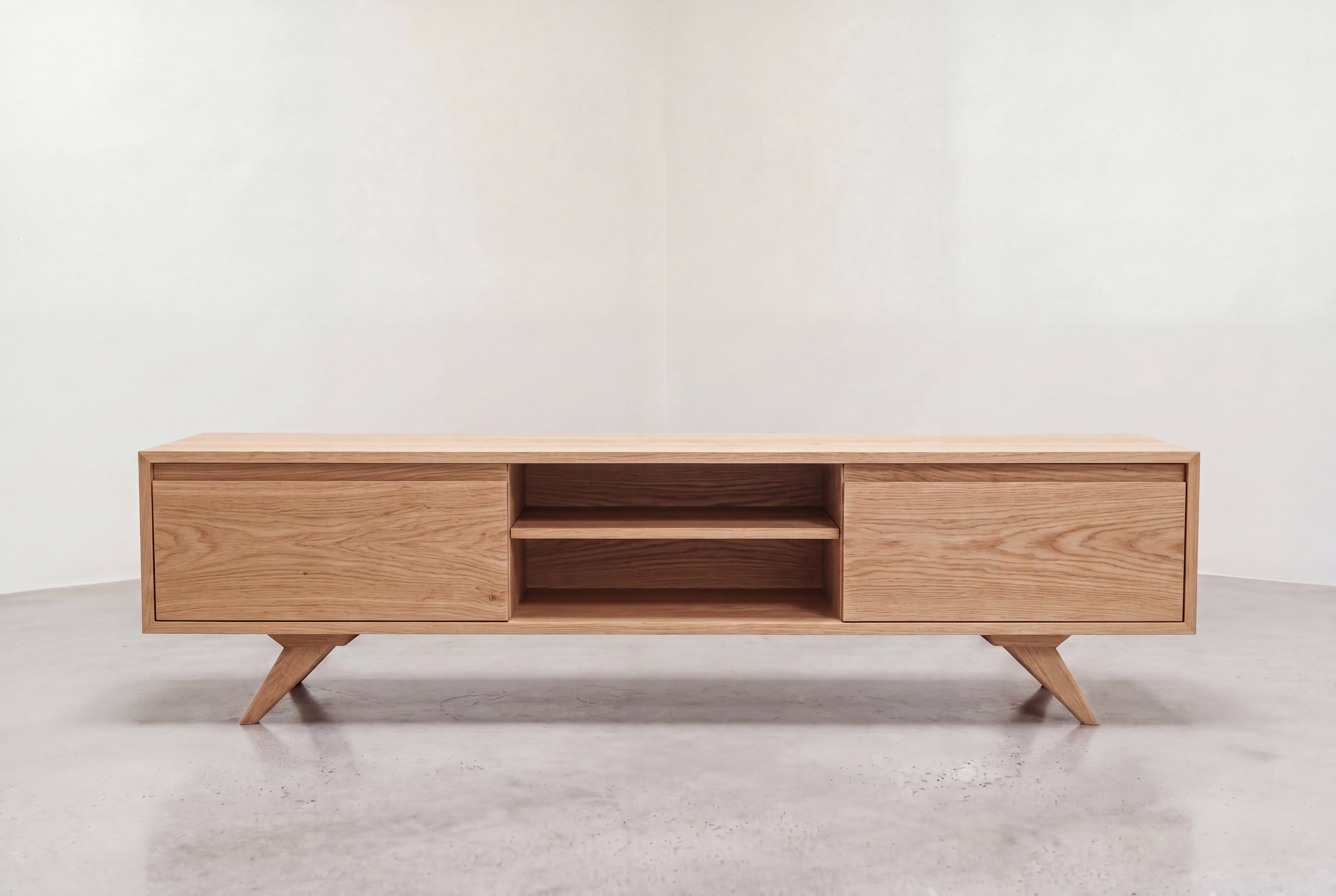The Modern Entertainment Console with Drawers - Solid Wood by TY Fine Furniture features a minimalist design with angled legs, two cabinet doors on each side, and two open shelves in the center for ample storage space. Perfectly positioned on a smooth, light gray floor against a plain wall, this contemporary piece serves as an ideal entertainment console for any living room.