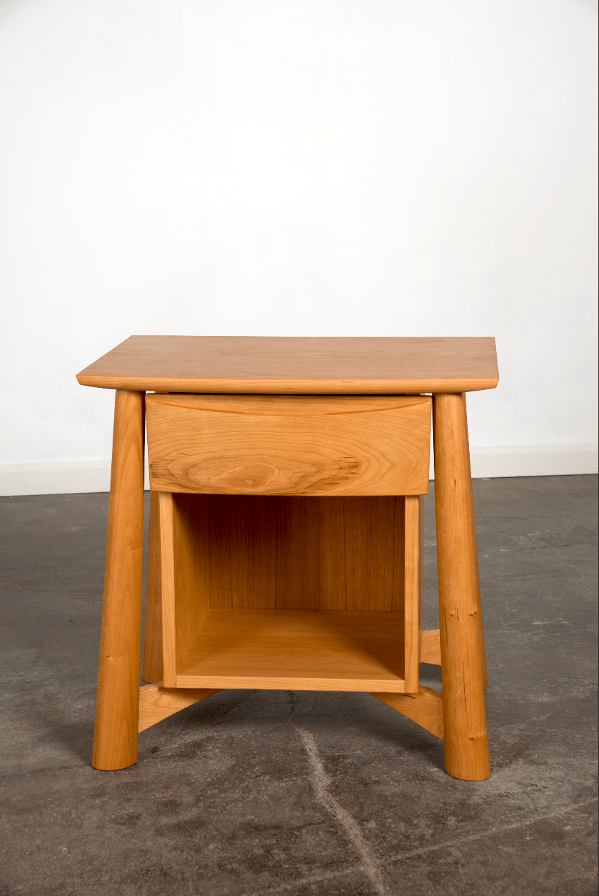 The Sunrise Side Table - Cherry by TY Fine Furniture is a Japanese-inspired solid wood table with a drawer, an open shelf below, and four round legs, shown on a concrete floor against a plain white wall.