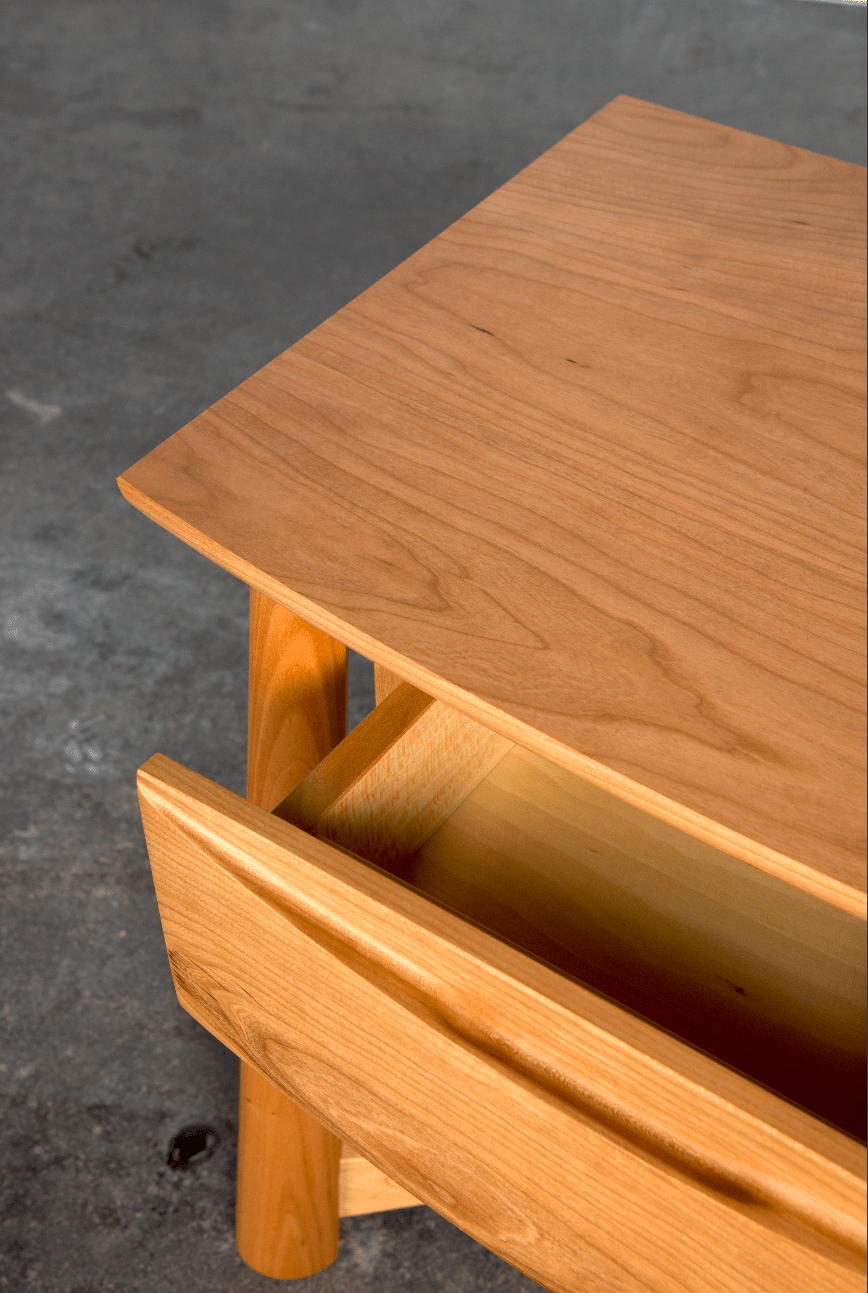 A close-up of the Sunrise Side Table - Cherry by TY Fine Furniture, featuring an open drawer, smooth light cherry wood grain, and rounded edges, displayed on a gray concrete floor.
