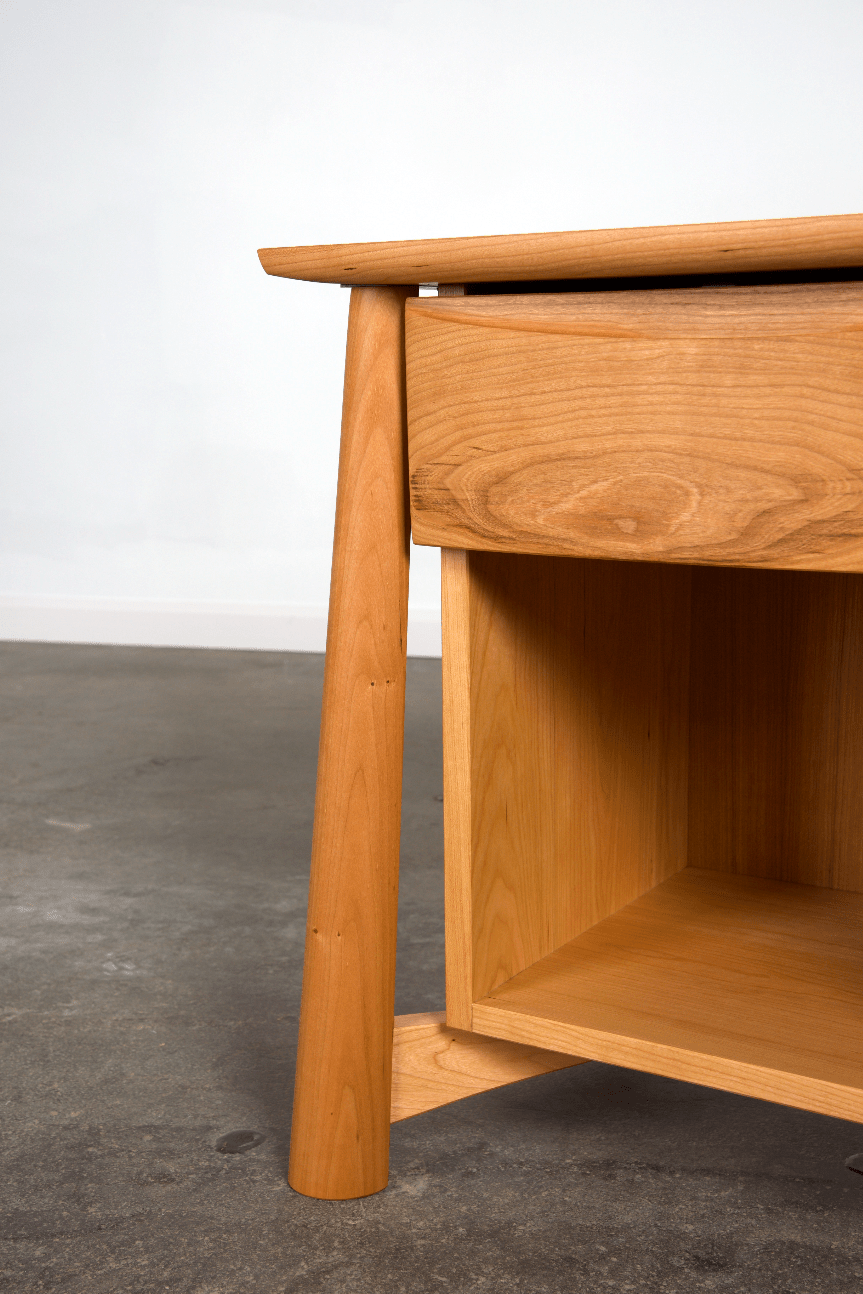 A close-up of the Sunrise Side Table - Cherry by TY Fine Furniture highlights its smooth, light cherry wood leg, a drawer above, and an open shelf below, set against a concrete floor and plain white wall.