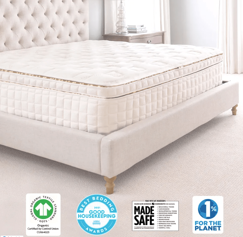 The Naturepedic EOS Organic Pillow Top Mattress, featuring a plush quilted surface, rests on a beige upholstered bed frame in a bright bedroom. Four badges—organic, award, safety, and environmental—appear at the bottom.