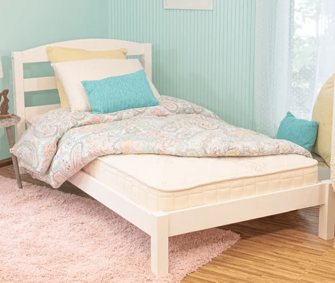 A cozy light blue room with wooden floors and a sheer-curtained window features a neatly made single bed with a white frame, topped with pastel paisley bedding, pillows, and the Naturepedic 2-in-1 Organic Kids Waterproof Mattress on a light pink rug.