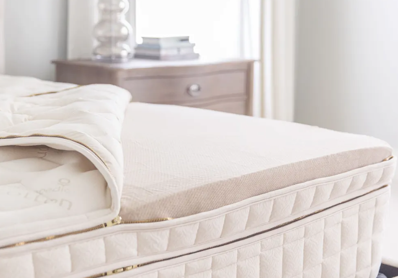 Close-up of a neatly made bed featuring the Naturepedic EOS Organic Pillow Top Mattress by Naturepedic, paired with a light wooden nightstand holding stacked books and a glass lamp, in a softly lit bedroom.