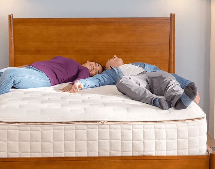 Two people relax on a Naturepedic EOS Classic Mattress with a wooden headboard, smiling in casual clothes on the white organic mattress, in a bright bedroom.