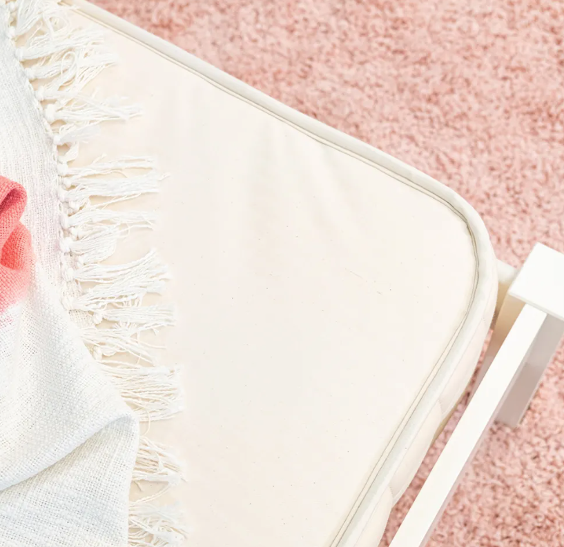 A close-up of a cream cushion on a white chair, draped with a white fringed blanket with pink accents, rests on a soft pink carpet beside the Naturepedic 2-in-1 Organic Kids Waterproof Mattress for a cozy, stylish touch.