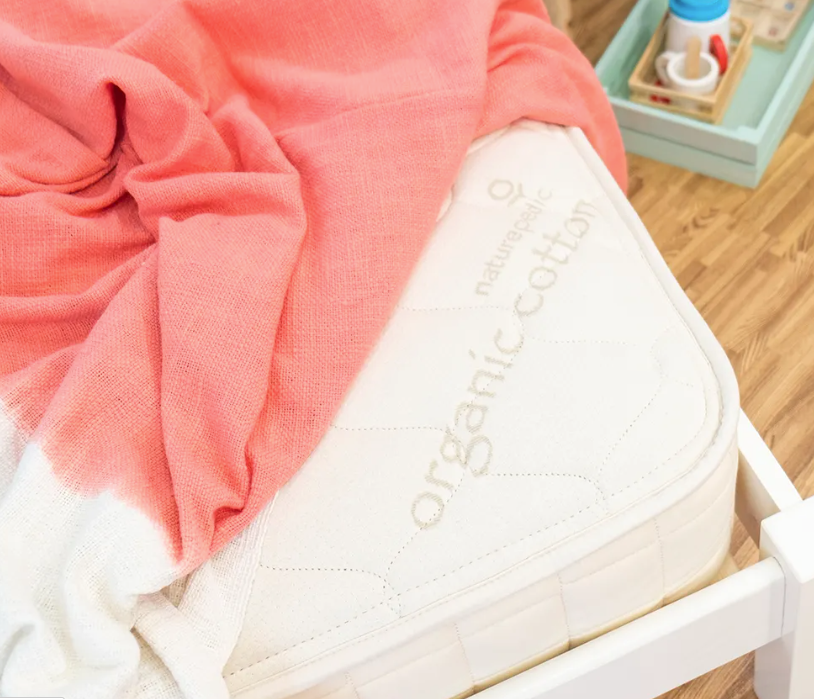 A close-up of the Naturepedic 2-in-1 Organic Kids Waterproof Mattress, GOTS certified and partially covered by a coral pink blanket, with a tray holding a baby bottle and items in the background on a wooden floor.