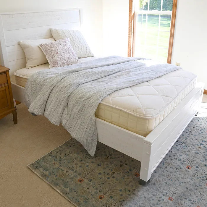 A sunlit room with beige carpet and a patterned blue rug features a Naturepedic Serenade Organic Hybrid Mattress by Naturepedic, half off the bed frame with an uncovered surface, a white headboard, white pillows, and a gray blanket.