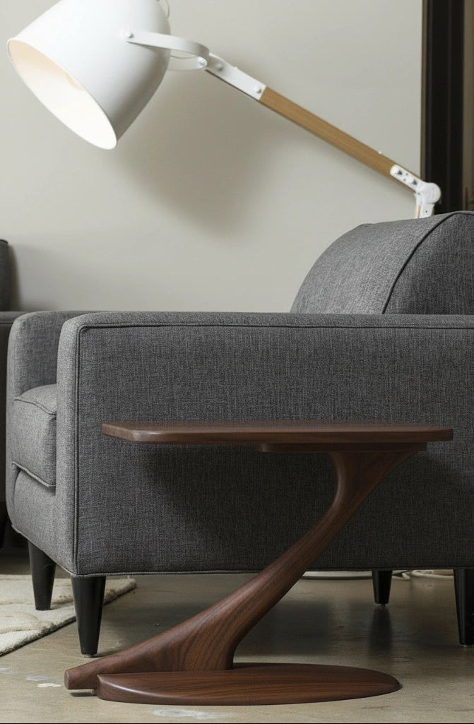 A modern living room features a gray upholstered sofa, the Cantilever Side Table Solid Wood Handmade - Walnut by TY Fine Furniture with its artisan curved base, and a large white floor lamp with a wooden arm angled over the sofa.