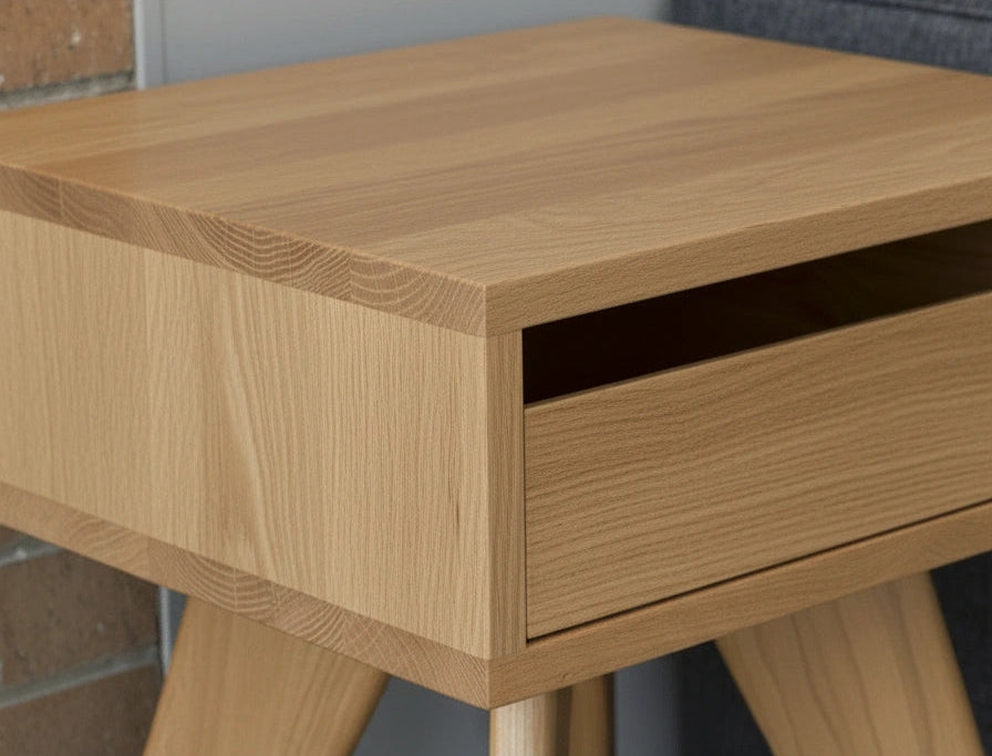 A close-up of the TY Fine Furniture Modern Apartment Side Table - White Oak, featuring angled legs, an open drawer, and solid white oak construction with a light wood grain finish, placed against a wall beside other furniture.