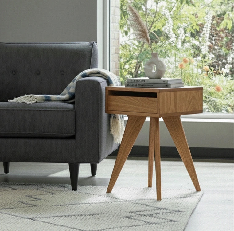 A modern living room with a gray sofa and a TY Fine Furniture Modern Apartment Side Table - White Oak, topped with a white vase of pampas grass and books. A window shows garden greenery; a patterned rug covers the floor.