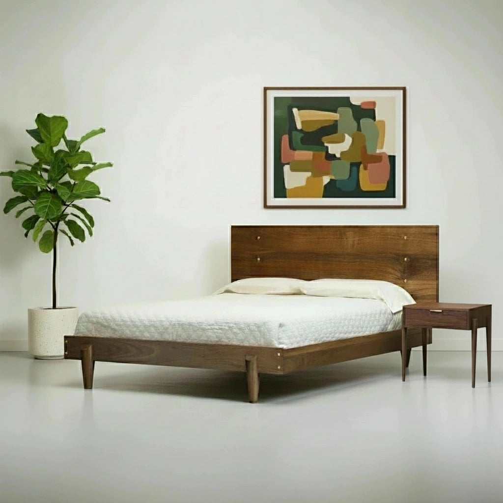 A modern bedroom showcases the TY Fine Furniture Apollo Platform Bed in solid walnut, topped with white bedding, paired with a wooden nightstand, a green potted plant, and an abstract colorful painting on a plain white wall.