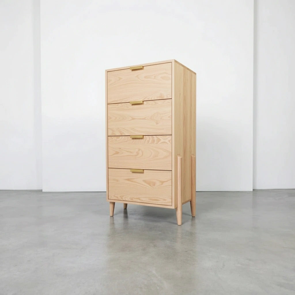 The Apollo Tall Dresser by TY Fine Furniture, crafted from solid wood with four drawers and minimalist brass handles, stands on tapered legs in a modern room with concrete floors and white walls.