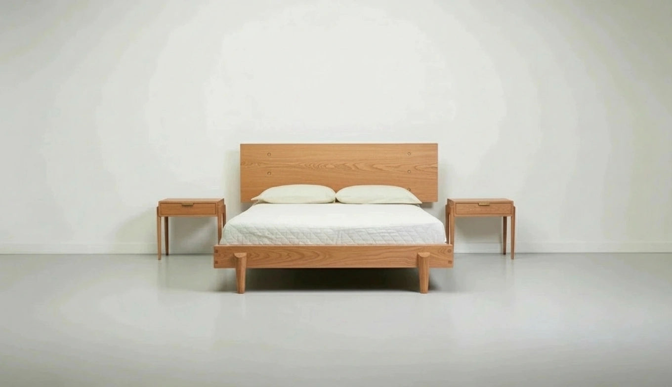 A minimalist bedroom featuring a wooden double bed, white pillows, a white quilt, and TY Fine Furniture's Apollo Solid Wood Side Tables—handcrafted in Columbus, Ohio from white oak—set against plain white walls and floor.
