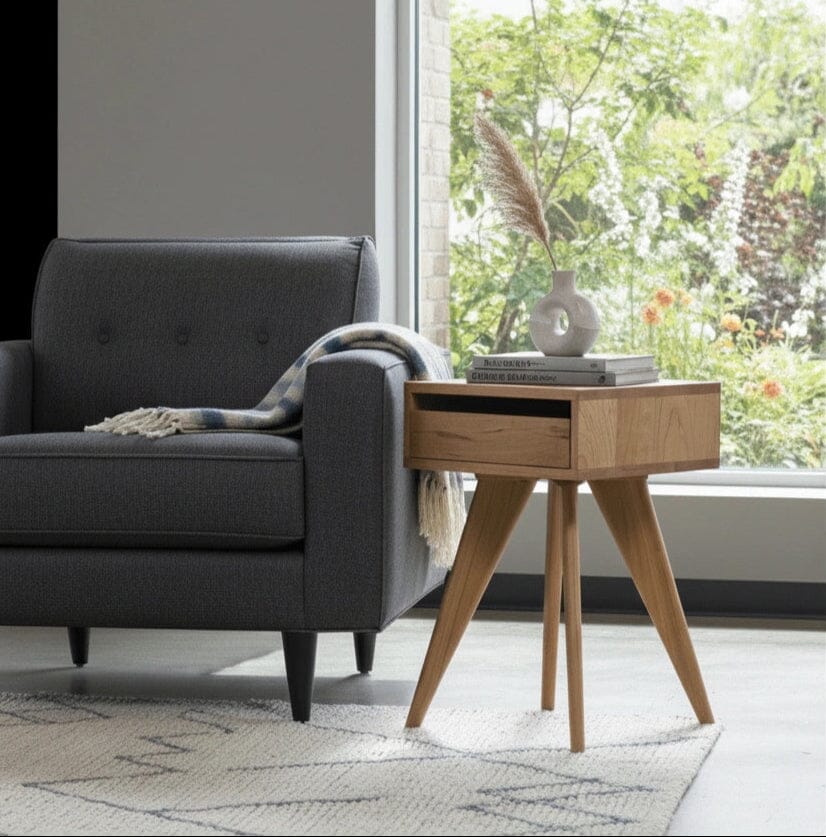 A modern living room corner features a dark grey armchair, plaid throw, and the TY Fine Furniture Modern Apartment Side Table in cherry, which holds books and a vase with dried grass beside a large window overlooking the garden.